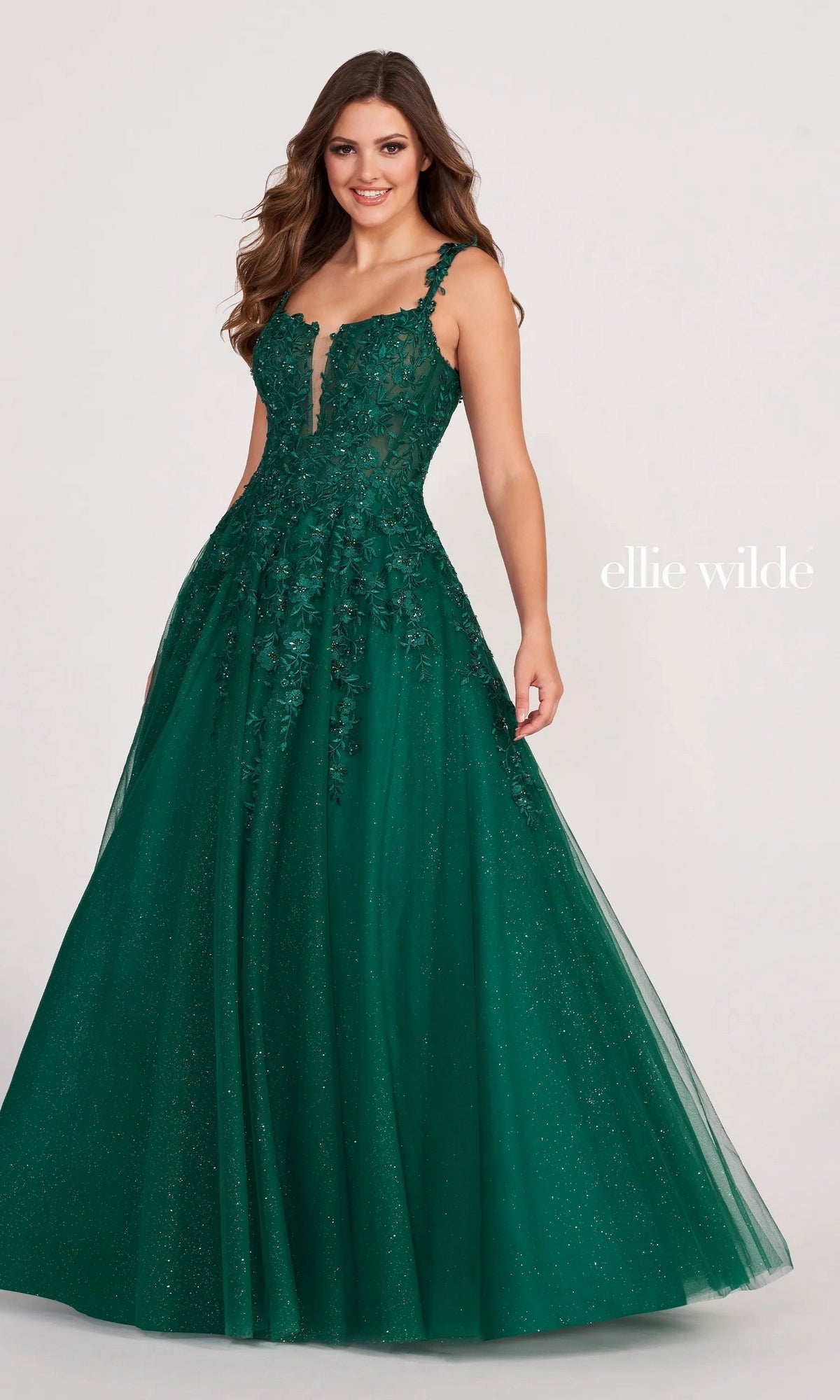 Woman wearing the Long Glitter Prom Ball Gown: Ellie Wilde EW120014, an emerald green floor-length A-line dress with a fitted bodice, floral appliqué, and glitter tulle details, standing against a plain background.